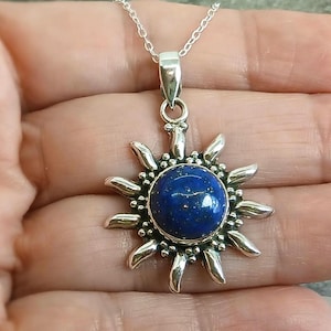 May include: A silver sun-shaped pendant featuring a round, deep blue lapis lazuli gemstone at its center. The pendant is suspended from a delicate silver chain, with metallic rays radiating outward from the stone, accented by small silver beads.