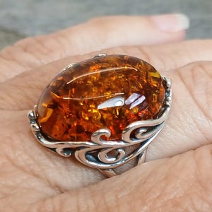 Op de afbeelding: Ovale barnsteen edelsteenring met een sterling zilveren filigraan zetting. De barnsteen steen is een warm, doorschijnend bruin met interne patronen. De zilveren zetting heeft een decoratief, wervelend ontwerp rond de steen.