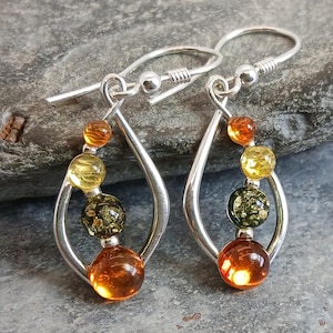 May include: A pair of silver drop earrings featuring a teardrop-shaped frame. Each earring is adorned with three spherical beads in varying shades of amber, including translucent orange, yellow, and dark green, suspended within the metal loop.