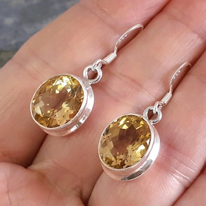 May include: A pair of silver earrings featuring oval-cut, golden-yellow gemstones. The gemstones are set in a bezel setting and suspended from a simple hook. The earrings are displayed against a neutral background, highlighting the jewelry's color and design.