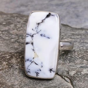 May include: A rectangular ring with a white stone featuring black veining. The stone is set in a silver band. The ring is displayed on a gray surface.