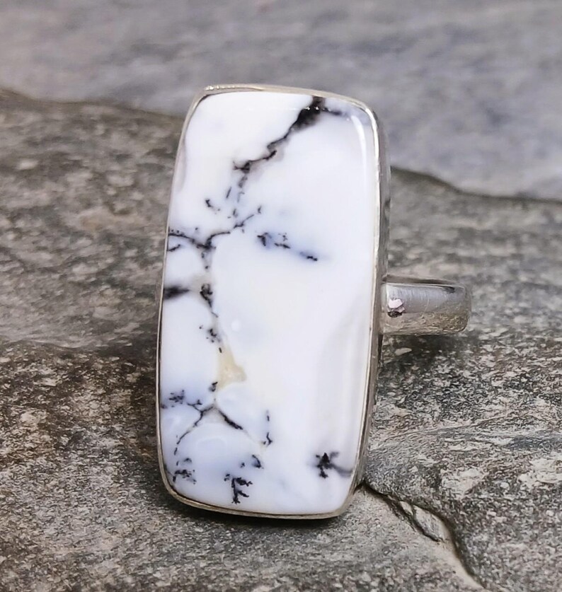 May include: A rectangular ring with a white stone featuring black veining. The stone is set in a silver band. The ring is displayed on a gray surface.