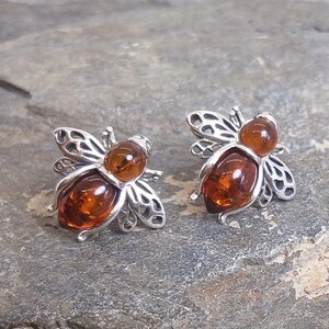 May include: A pair of bee-shaped stud earrings crafted from silver-toned metal. Each earring features a smooth, rounded, translucent amber-colored stone for the head and a larger, teardrop-shaped amber-colored stone for the body, with openwork wings.