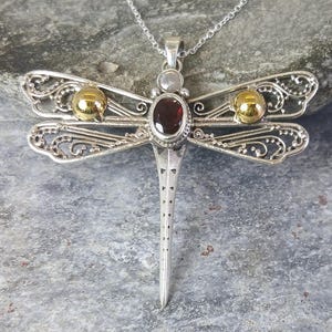 May include: A silver dragonfly pendant necklace featuring intricate filigree wings, a central oval deep red garnet gemstone, and two small polished gold spheres on the upper wings. The pendant hangs from a delicate silver chain.