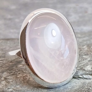 Puede incluir: Un anillo de plata con una gema grande, ovalada y rosa pálido. La gema tiene una superficie lisa y pulida con un brillo sutil y nacarado. La banda y el engaste del anillo son de metal plateado brillante.