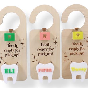 May include: Three wooden door hangers for the tooth fairy, each with a unique design. Each hanger has a small bottle, a tooth-shaped cutout with a name, and the text "Tooth ready for pick up!". The names are Eli, Piper, and Davey.