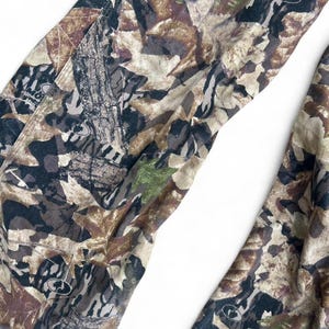 May include: Pants featuring a camouflage pattern of brown, tan, black, and green leaves and tree bark textures. The fabric includes small circular logos with the text Mossy Oak printed throughout the design in a repeating, detailed forest motif.