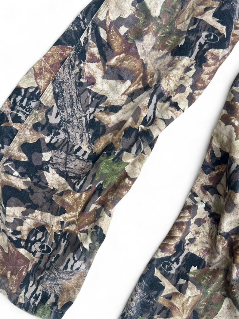 May include: Pants featuring a camouflage pattern of brown, tan, black, and green leaves and tree bark textures. The fabric includes small circular logos with the text Mossy Oak printed throughout the design in a repeating, detailed forest motif.