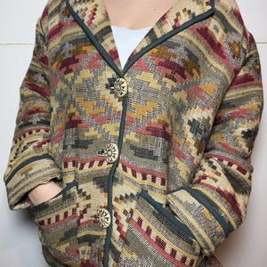 May include: A patterned jacket with a geometric design in shades of beige, red, and green. The jacket has a collar, pockets, and large decorative buttons. The trim is a dark green color.