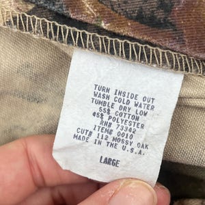 May include: A white fabric garment care label with black text reading: TURN INSIDE OUT, WASH COLD WATER, TUMBLE DRY LOW, 55% COTTON, 45% POLYESTER, RN# 73342, ITEM# 0010, CUT# 112 MOSSY OAK, MADE IN THE U.S.A., LARGE. The label is attached to camouflage fabric.