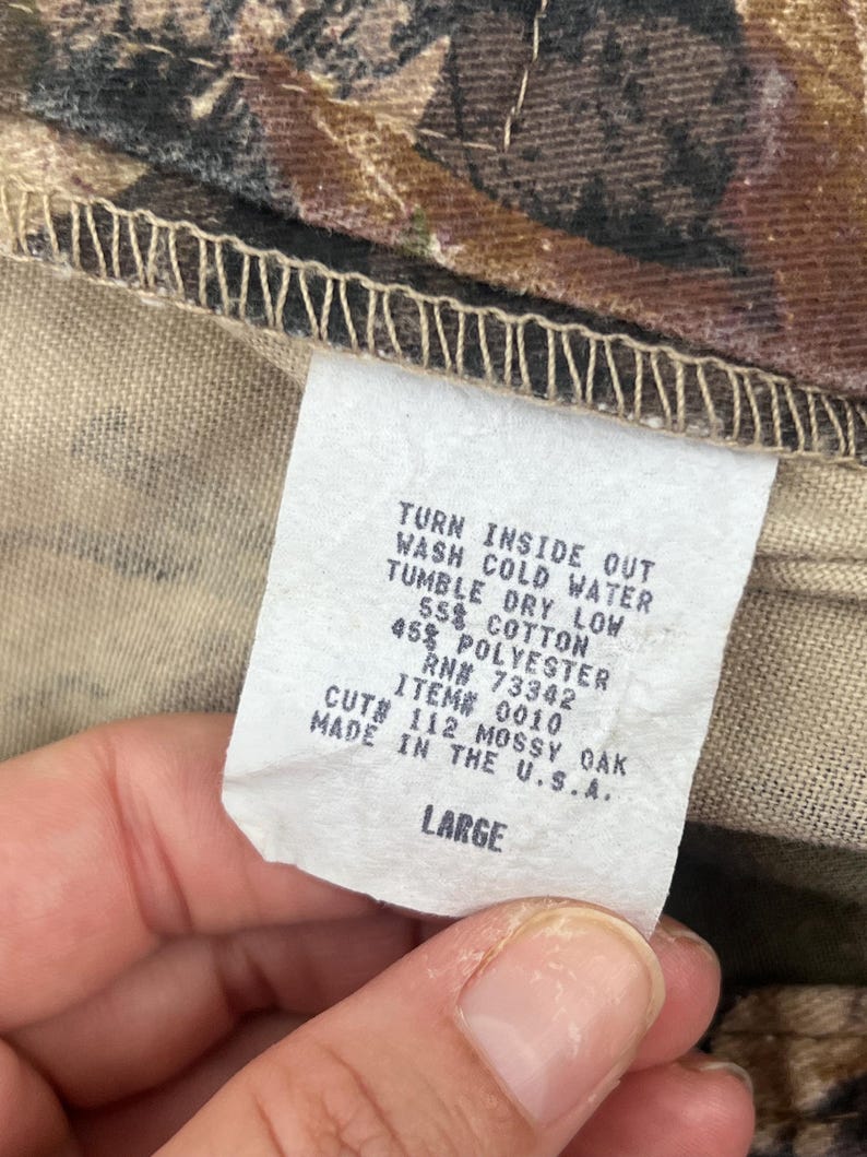 May include: A white fabric garment care label with black text reading: TURN INSIDE OUT, WASH COLD WATER, TUMBLE DRY LOW, 55% COTTON, 45% POLYESTER, RN# 73342, ITEM# 0010, CUT# 112 MOSSY OAK, MADE IN THE U.S.A., LARGE. The label is attached to camouflage fabric.