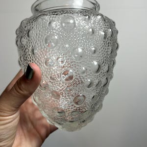 May include: Clear glass jar with a textured surface of small and large bubbles. The jar has a wide opening and a rounded body. The glass is transparent, allowing light to pass through. The jar is held by a hand.