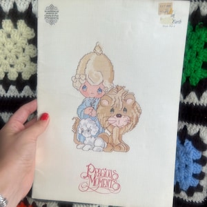May include: A white cross-stitch pattern booklet titled Precious Moments featuring a graphic of a child with blonde hair in a blue outfit sitting next to a brown lion and a small white lamb. The cover includes the text Designs by Gloria & Pat.