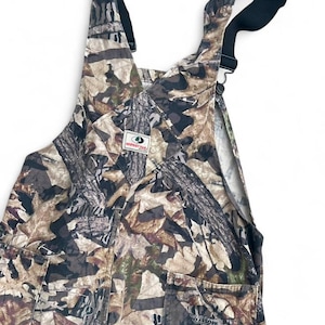 May include: Mossy Oak camouflage overalls featuring a brown, tan, and black leaf and tree bark pattern. The garment includes adjustable black shoulder straps with metal hardware, a front bib pocket with a Mossy Oak brand patch, and two side pockets.
