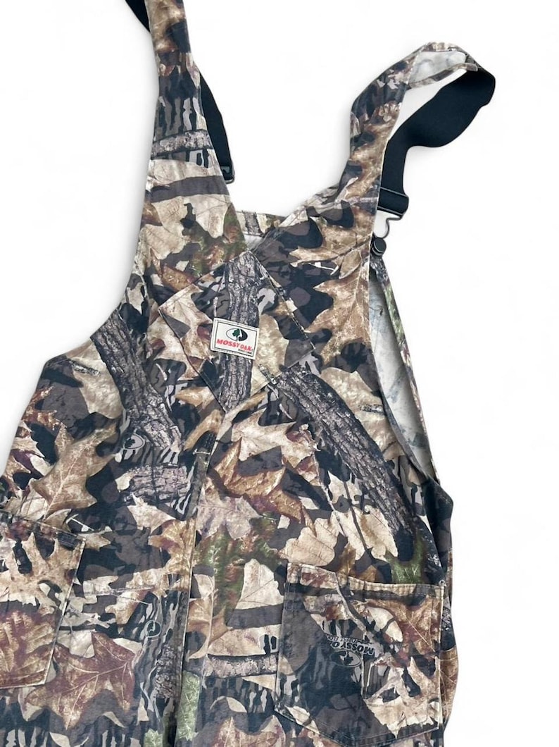 May include: Mossy Oak camouflage overalls featuring a brown, tan, and black leaf and tree bark pattern. The garment includes adjustable black shoulder straps with metal hardware, a front bib pocket with a Mossy Oak brand patch, and two side pockets.