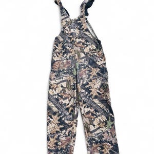 May include: Full-length overalls featuring a camouflage pattern of brown, tan, and dark green leaves and branches. The garment includes adjustable shoulder straps with black plastic buckles and a central chest pocket with a small white brand label.