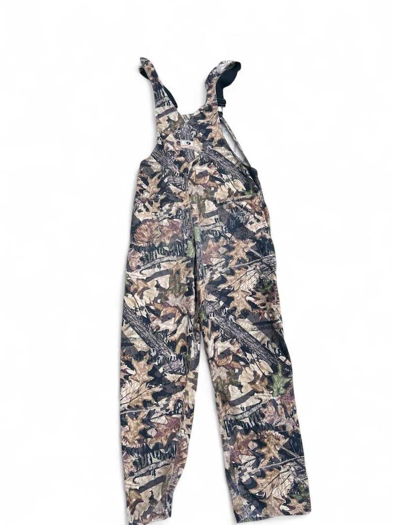 May include: Full-length overalls featuring a camouflage pattern of brown, tan, and dark green leaves and branches. The garment includes adjustable shoulder straps with black plastic buckles and a central chest pocket with a small white brand label.