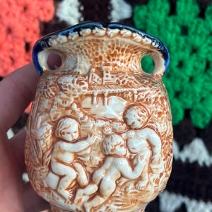 May include: A small ceramic vase featuring a relief design of three figures in a landscape. The piece has a cream-colored body with brown detailing and a dark blue rim and base. It includes two small circular handles at the top opening.