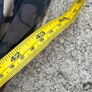 May include: A piece of fabric with a dark brown, black, and tan camouflage pattern featuring visible stitching along the edge. A yellow measuring tape is placed alongside the fabric, showing a measurement between 42 and 45 inches in length.