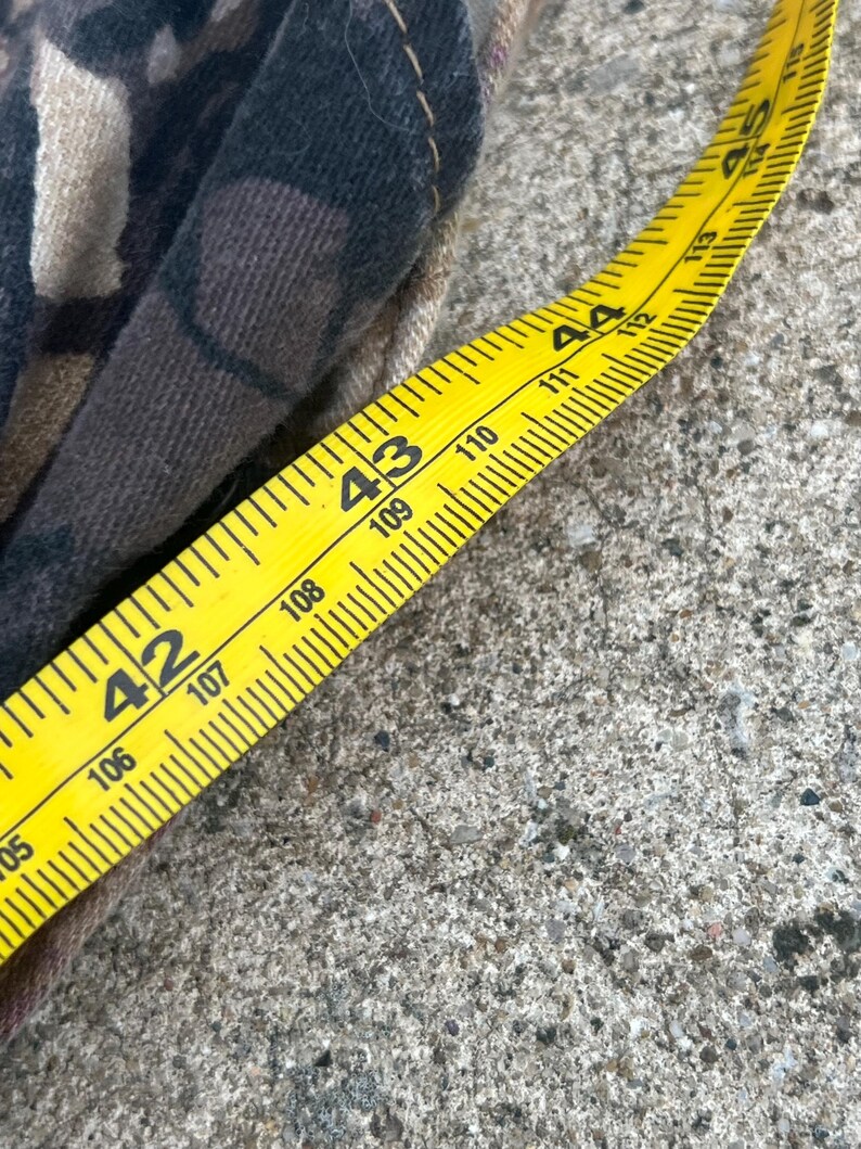 May include: A piece of fabric with a dark brown, black, and tan camouflage pattern featuring visible stitching along the edge. A yellow measuring tape is placed alongside the fabric, showing a measurement between 42 and 45 inches in length.