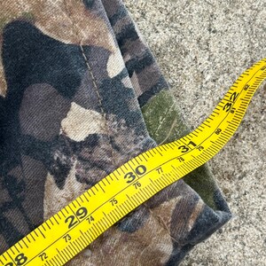 May include: A pair of camouflage pants featuring a brown, black, and green woodland pattern. A yellow measuring tape is placed across the fabric, indicating a waist measurement of approximately 32 inches along the top edge of the garment.