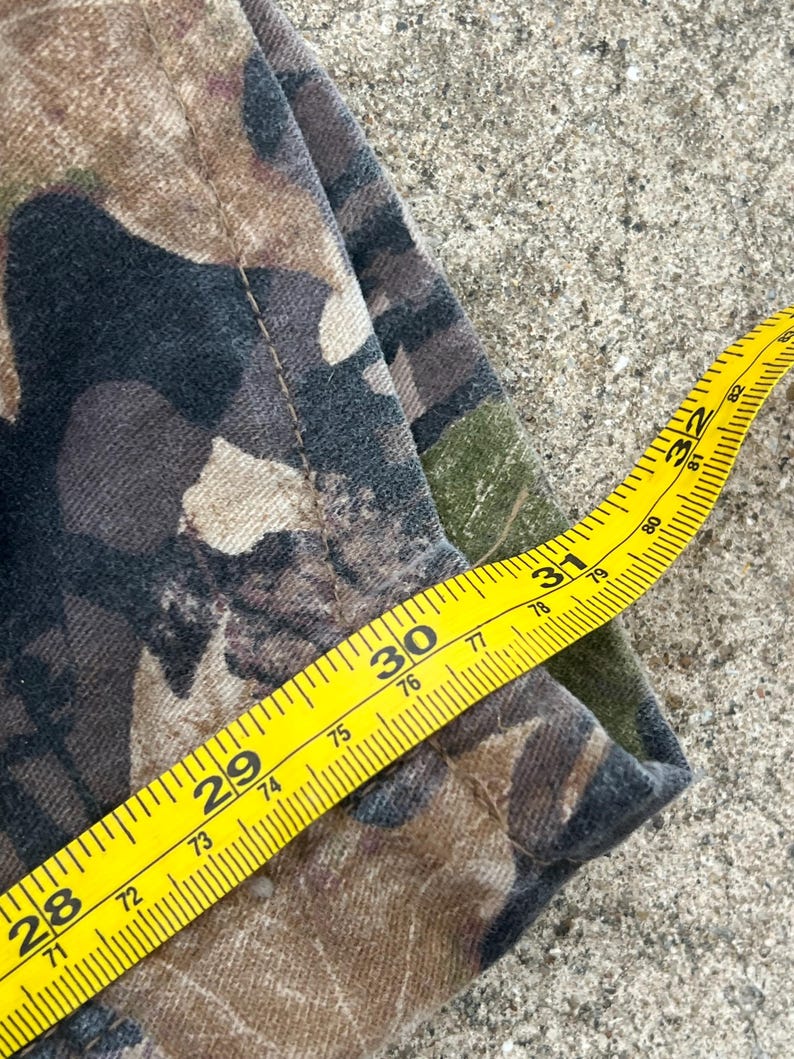 May include: A pair of camouflage pants featuring a brown, black, and green woodland pattern. A yellow measuring tape is placed across the fabric, indicating a waist measurement of approximately 32 inches along the top edge of the garment.