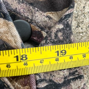 May include: A camouflage patterned garment featuring a black button closure. A yellow measuring tape is positioned across the fabric, showing a measurement between 18 and 19 inches, with metric markings indicating approximately 46 to 49 centimeters.