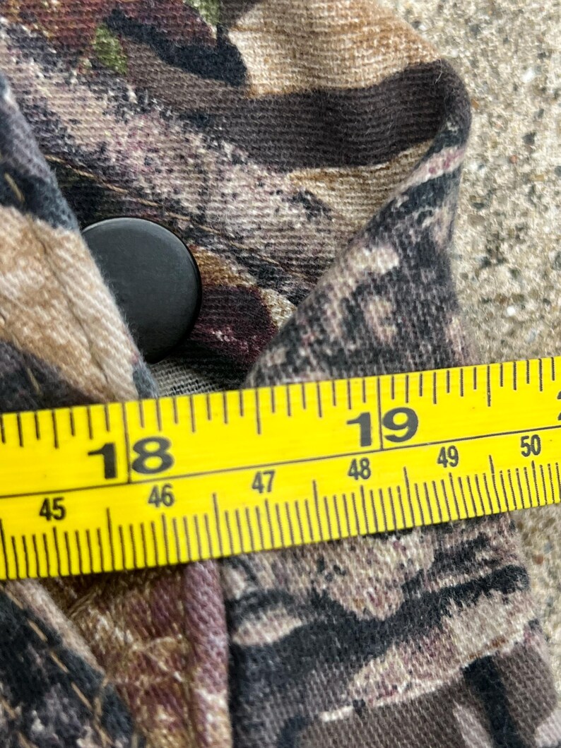 May include: A camouflage patterned garment featuring a black button closure. A yellow measuring tape is positioned across the fabric, showing a measurement between 18 and 19 inches, with metric markings indicating approximately 46 to 49 centimeters.