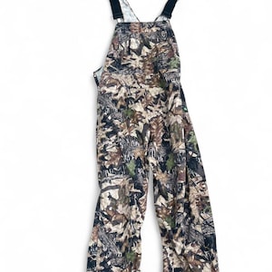 May include: Full-length overalls featuring a detailed forest camouflage pattern in shades of brown, tan, green, and black. The garment includes a chest pocket, adjustable black shoulder straps with metal hardware, and a straight-leg design.