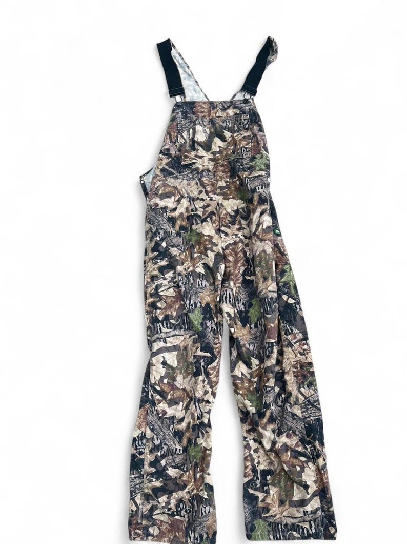 May include: Full-length overalls featuring a detailed forest camouflage pattern in shades of brown, tan, green, and black. The garment includes a chest pocket, adjustable black shoulder straps with metal hardware, and a straight-leg design.