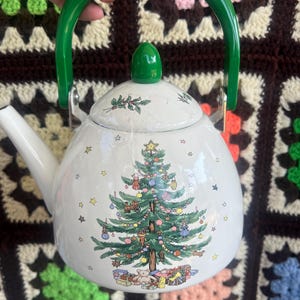 May include: A white tea kettle featuring a green handle and a matching green lid knob. The body of the kettle is decorated with a detailed illustration of a Christmas tree adorned with ornaments, surrounded by small stars and gifts at the base.