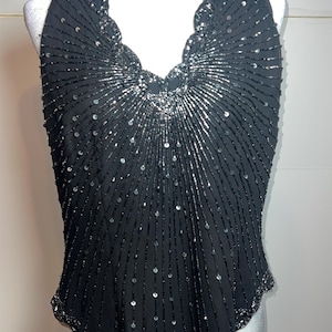 May include: Black halter top with silver bead and sequin embellishments. The top features a scalloped neckline and hemline, with vertical beaded lines radiating from the center. The design includes a V-neck and a sleeveless style, creating a glamorous look.