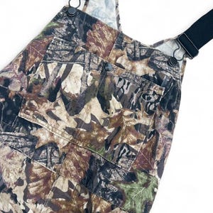 May include: Overalls featuring a brown, green, and tan leaf camouflage pattern. The garment includes black elastic shoulder straps with metal hardware, a large front bib pocket with a snap closure, and a small logo patch on the pocket.