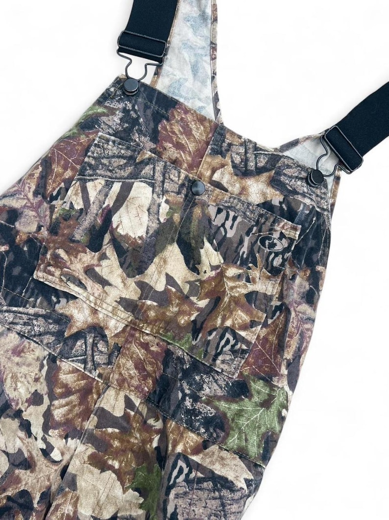 May include: Overalls featuring a brown, green, and tan leaf camouflage pattern. The garment includes black elastic shoulder straps with metal hardware, a large front bib pocket with a snap closure, and a small logo patch on the pocket.