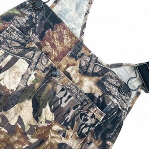 May include: Overalls featuring a camouflage pattern of brown, tan, and black leaves and branches. The garment includes black adjustable shoulder straps with metal hardware, a front chest pocket with a small circular logo, and black buttons.