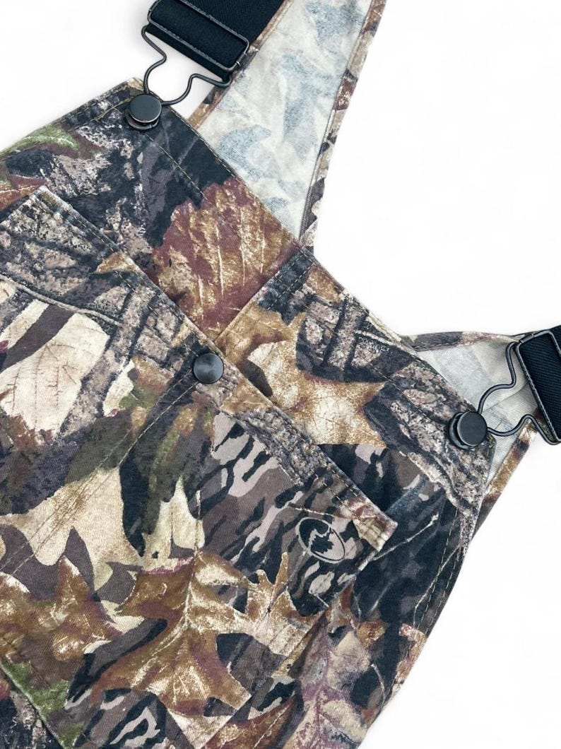 May include: Overalls featuring a camouflage pattern of brown, tan, and black leaves and branches. The garment includes black adjustable shoulder straps with metal hardware, a front chest pocket with a small circular logo, and black buttons.