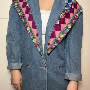 May include: A light blue denim blazer with a unique patchwork collar. The collar features a geometric pattern of pink and purple squares, accented with floral trim. The blazer has a single button closure and cuffed sleeves.