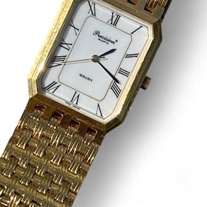 May include: A gold-toned Gruen Precision Quartz watch with a rectangular face and a woven metal band. The watch face is white with black Roman numerals and hands. The watch is a vintage style.