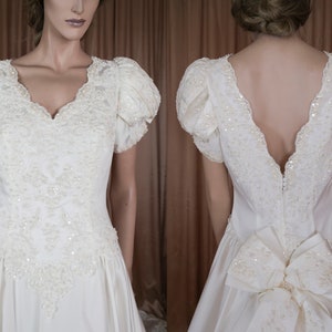 May include: A white satin wedding dress with a V-neckline and short puffed sleeves. The dress is embellished with pearl and sequin beading and features a large bow at the back.