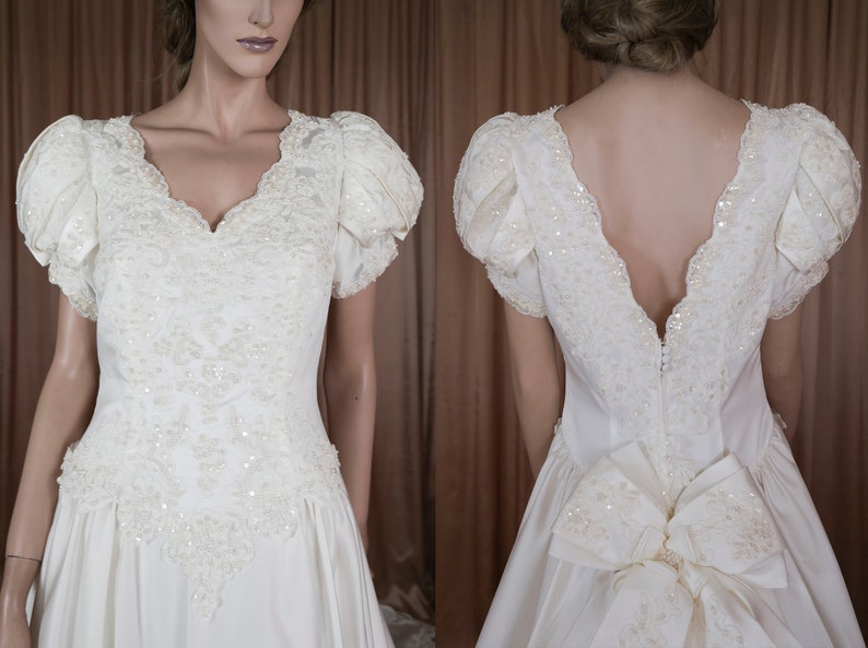 May include: A white satin wedding dress with a V-neckline and short puffed sleeves. The dress is embellished with pearl and sequin beading and features a large bow at the back.