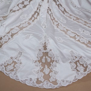 May include: White satin wedding dress with a lace overlay. The lace is a delicate floral pattern with a scalloped edge.