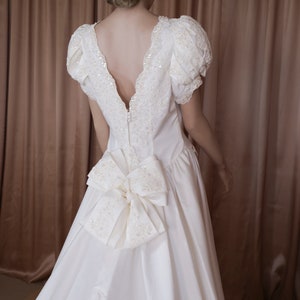 May include: A white satin wedding dress with a low back and a large bow at the back. The dress has a lace trim and is decorated with sequins.