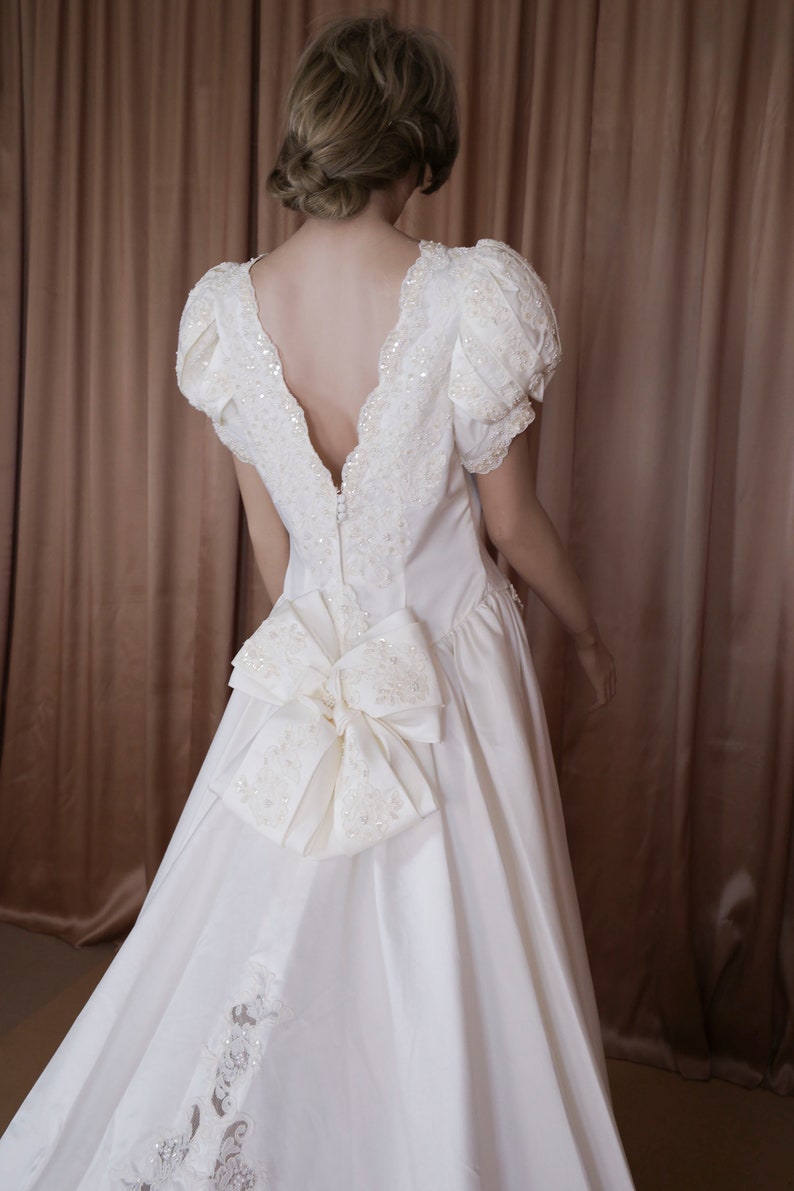 May include: A white satin wedding dress with a low back and a large bow at the back. The dress has a lace trim and is decorated with sequins.