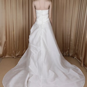 May include: A white, strapless wedding dress with a long train. The back of the gown has a button closure and a fitted bodice. The dress is made of a flowing, lightweight fabric.