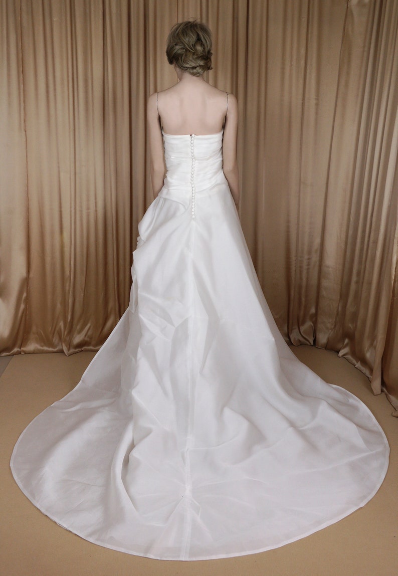 May include: A white, strapless wedding dress with a long train. The back of the gown has a button closure and a fitted bodice. The dress is made of a flowing, lightweight fabric.