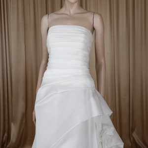 May include: A white strapless wedding dress on a mannequin. The bodice is ruched, and the skirt has layered fabric with a floral accent. The dress has a flowing, floor-length design, suitable for bridal wear.