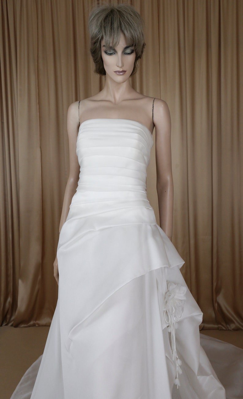 May include: A white strapless wedding dress on a mannequin. The bodice is ruched, and the skirt has layered fabric with a floral accent. The dress has a flowing, floor-length design, suitable for bridal wear.