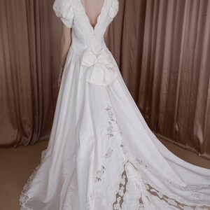 May include: A white satin wedding dress with a lace overlay and a large bow at the back. The dress has a deep V-neckline and short sleeves.
