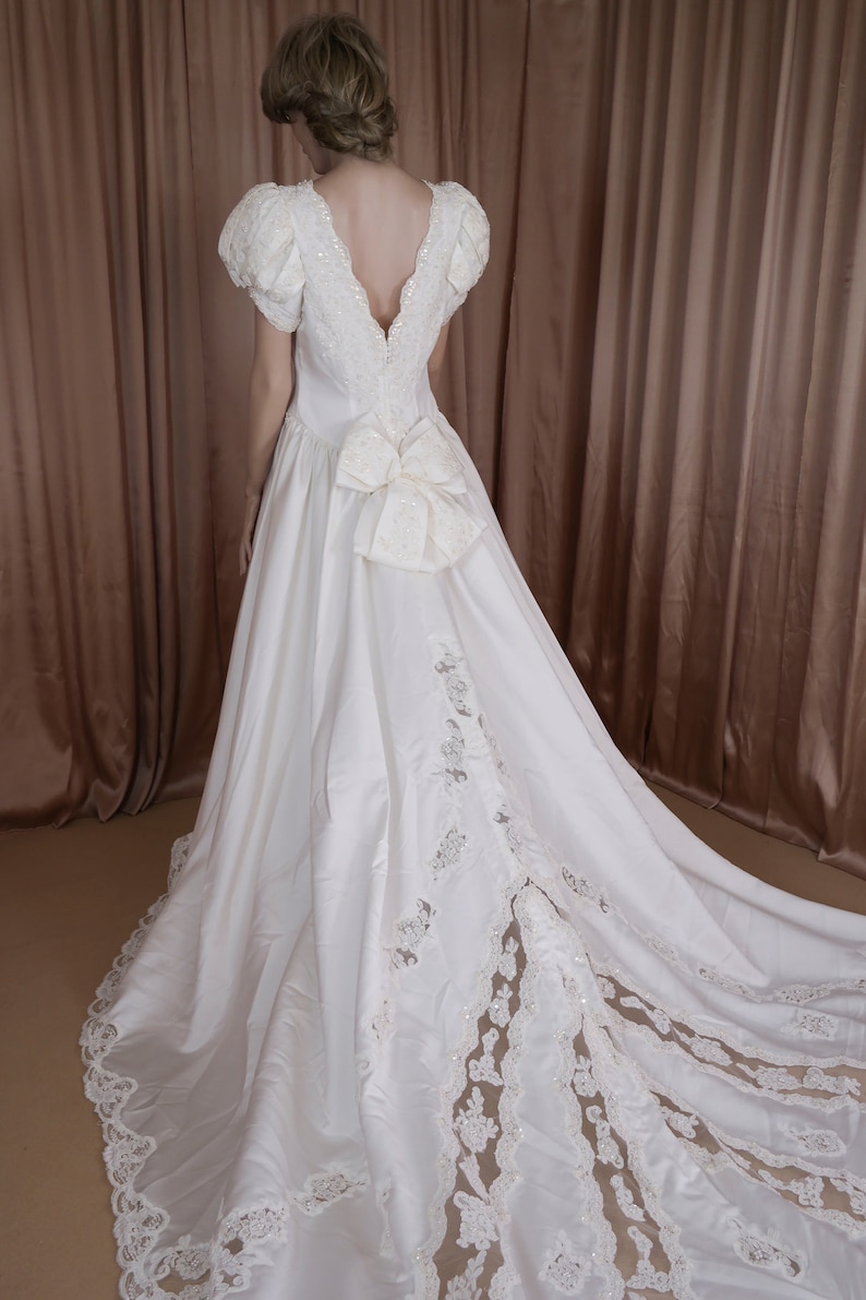 May include: A white satin wedding dress with a lace overlay and a large bow at the back. The dress has a deep V-neckline and short sleeves.