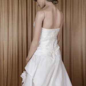May include: A white bridal gown with a fitted bodice and a flowing skirt. The back of the dress has a row of small buttons. The dress features thin straps and a layered skirt detail. The mannequin is positioned in front of a gold curtain.